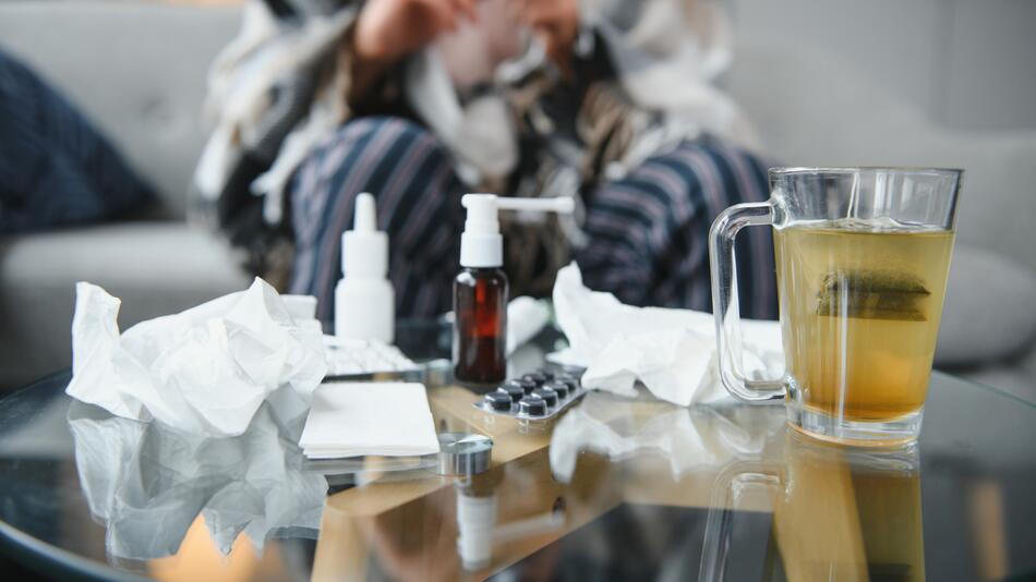 Medikamente, Tee und Nasenspray stehen auf einem Tisch, eine Person sitzt krank auf der Couch