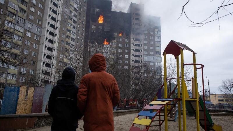 Zwei Menschen stehen in der Ukraine vor einem brennenden Gebäude.