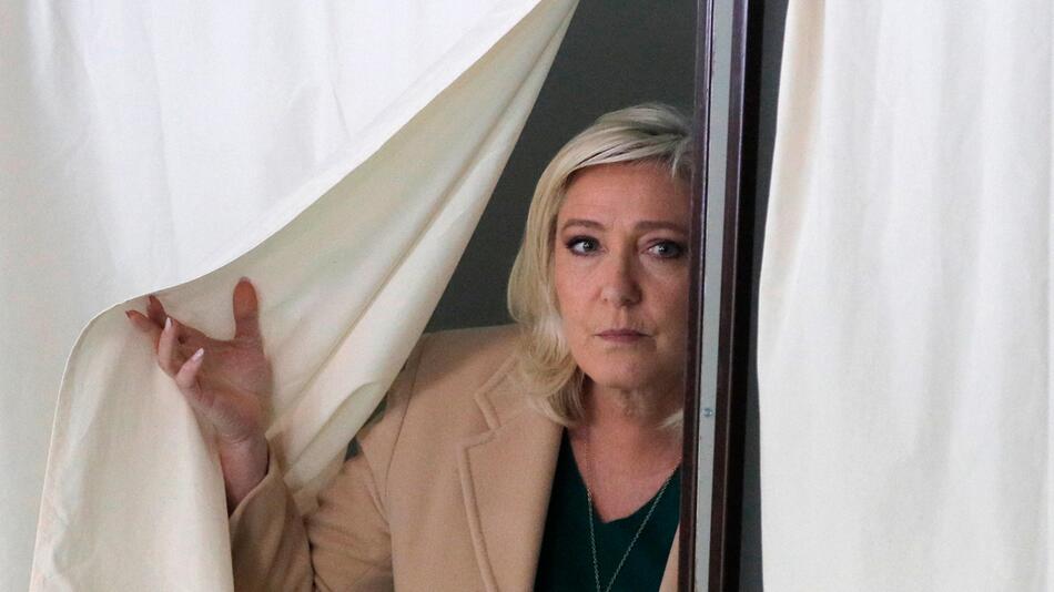 Portrait von Marine Le Pen, die den Vorhang einer Wahlkabine zur Seite schiebt.