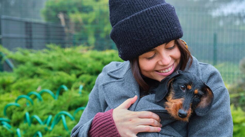 Eine Frau lächelt einen Hund an, der unter ihrem Mantel eingekuschelt ist.