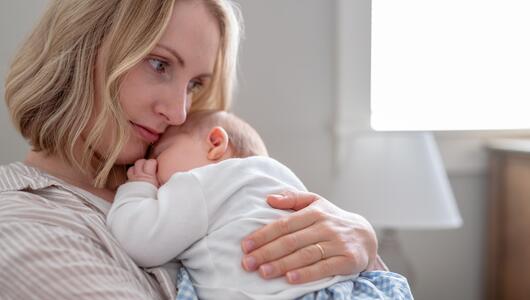 Eine Mutter hält ihren schlafenden Säugling im Arm, sie wirkt nachdenklich.