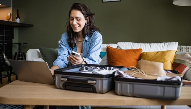 Frau sitzt mit Laptop und Smartphone vor ihrem Koffer.