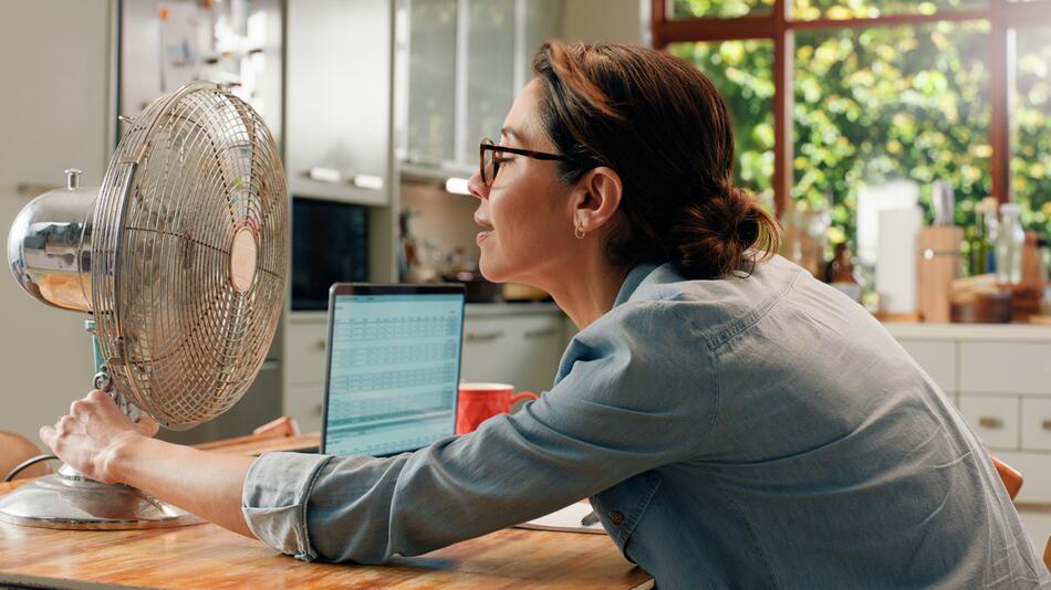 Frau sitzt Zuhause und arbeitet am Laptop, sie kühlt sich mit einem Ventilator