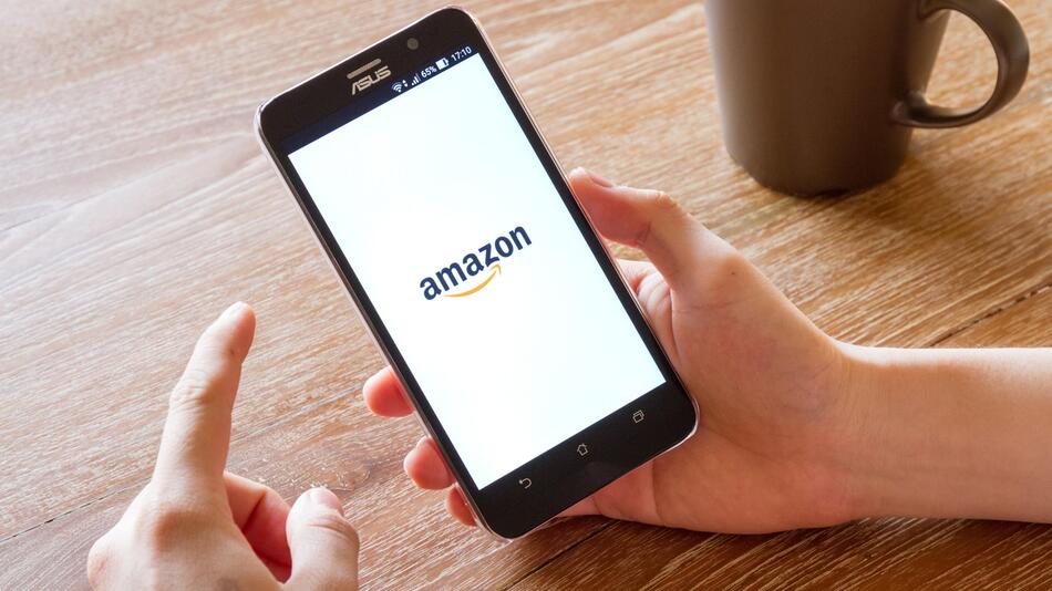 Ein Mensch hält ein Smartphone in der Hand, auf dem Bildschirm ist das Logo von Amazon zu sehen.