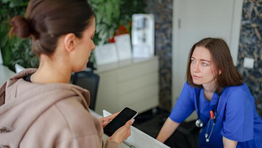 Frau, die auf Handy schaut - am Empfang einer Arztpraxis. Die Mitarbeiterin schaut genervt. 
