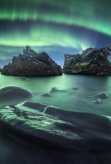 Starke Polarlichter bedecken den Himmel über Felsen und dem Meer