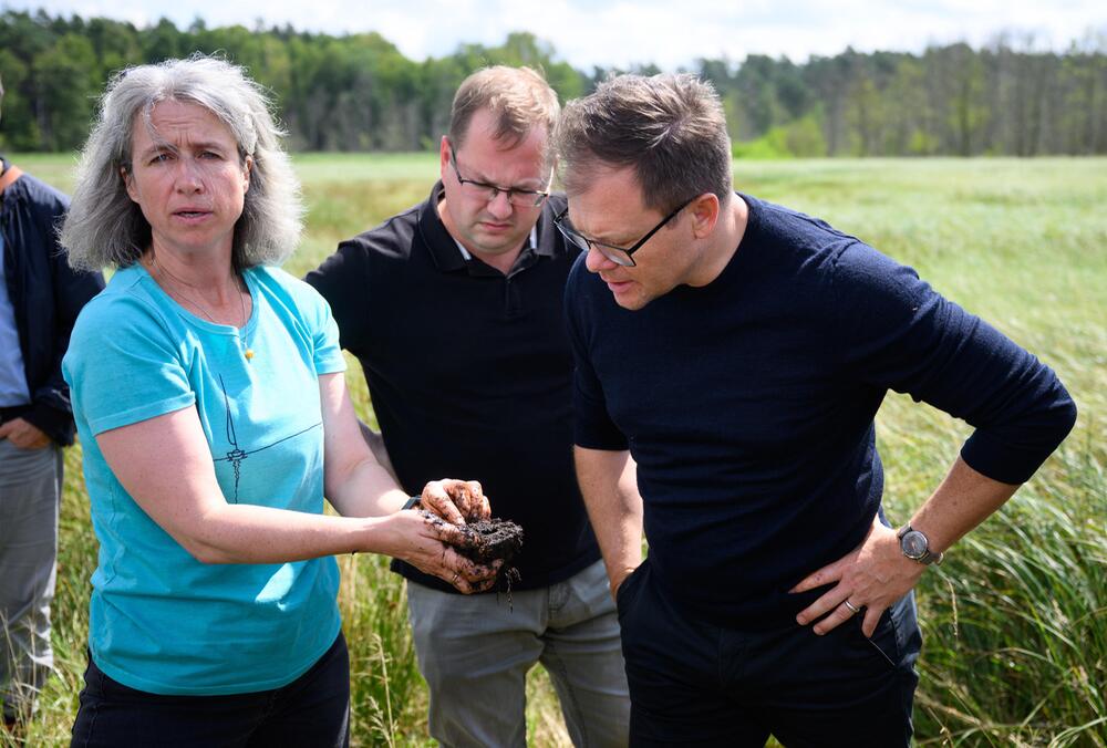 Bundesumweltminister Schneider schaut Torf an, den Anje Marten hält. Mit Landwirt Fischer.